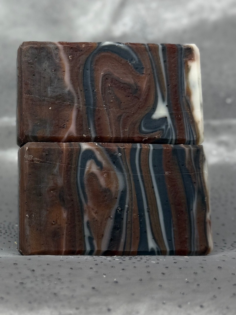 Two stacked Coffee, Cocoa, & Charcoal soap bars with brown, black and white wood-grain swirls, on grey satin