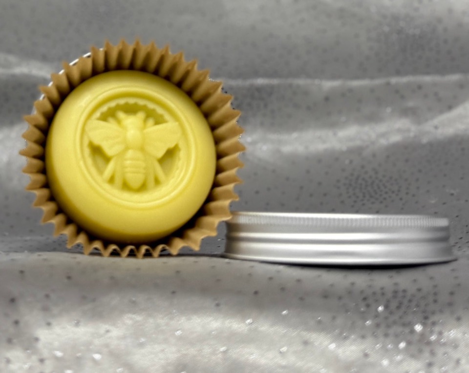 Round pale yellow Lotion Bar embossed with a bee, in a brown paper cup, beside a silver tin lid on grey satin